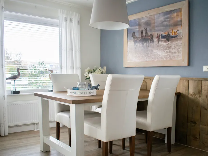 De eethoek: een tafel met witte stoelen onder een grote witte lamp, en op de vensterbank twee vogelbeeldjes met lange snavels.
