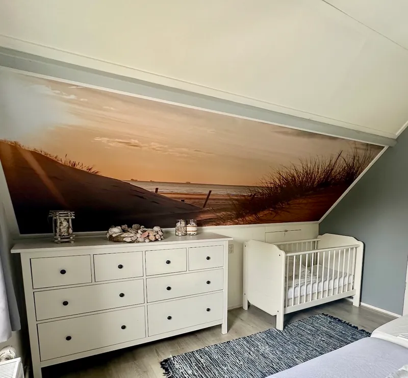 De slaapkamer met het tweepersoonsbed: een wit kinderbedje en een ladekast met schelpen, voor een fotobehang van duin en zee.