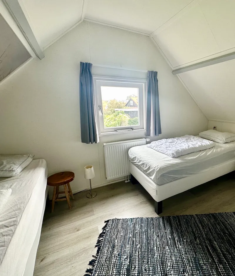 De slaapkamer met twee eenpersoonsbedden: een houten krukje met een lampje ernaast, en een klein raam met blauwe gordijnen.