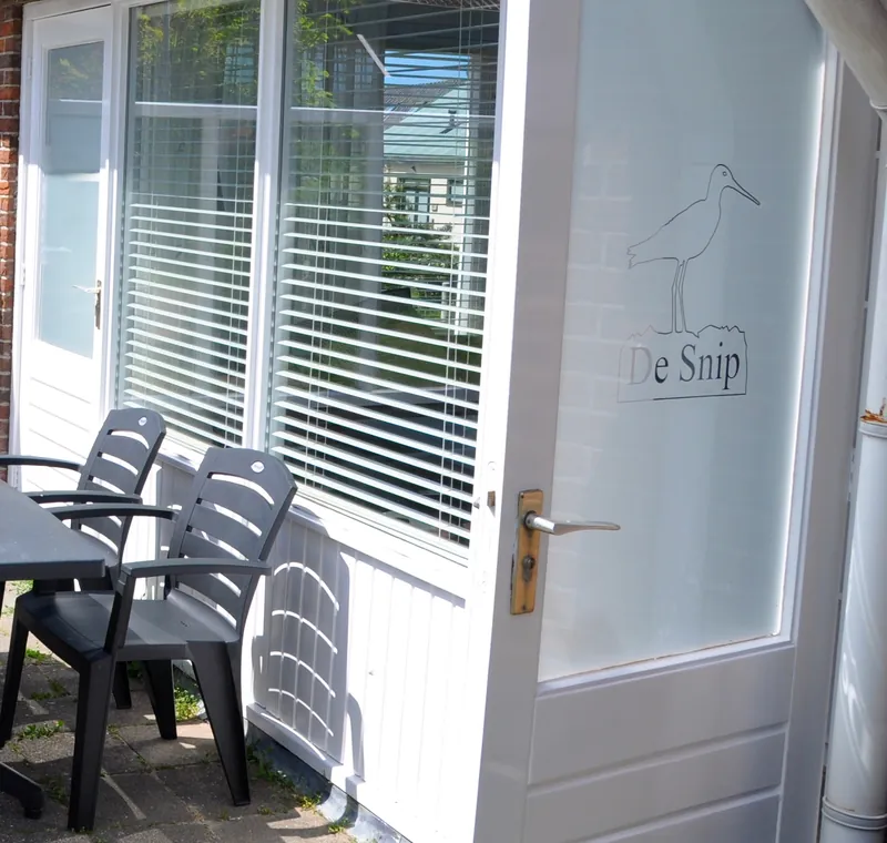 Het terras: tafel en stoelen in de zon, en de deur met de naam De Snip en een getekende vogel met een lange snavel op het glas.