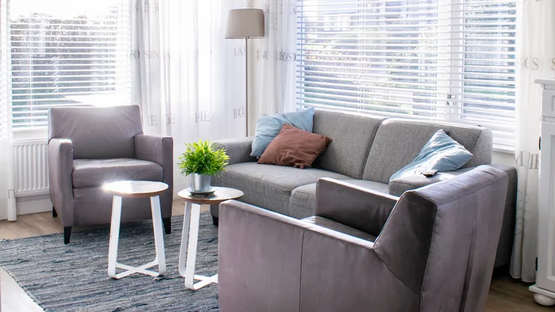 De woonkamer: een grijze bank met blauwe en bruine kussens, twee grijze fauteuils, twee ronde tafeltjes en een blauwgrijs vloerkleed.