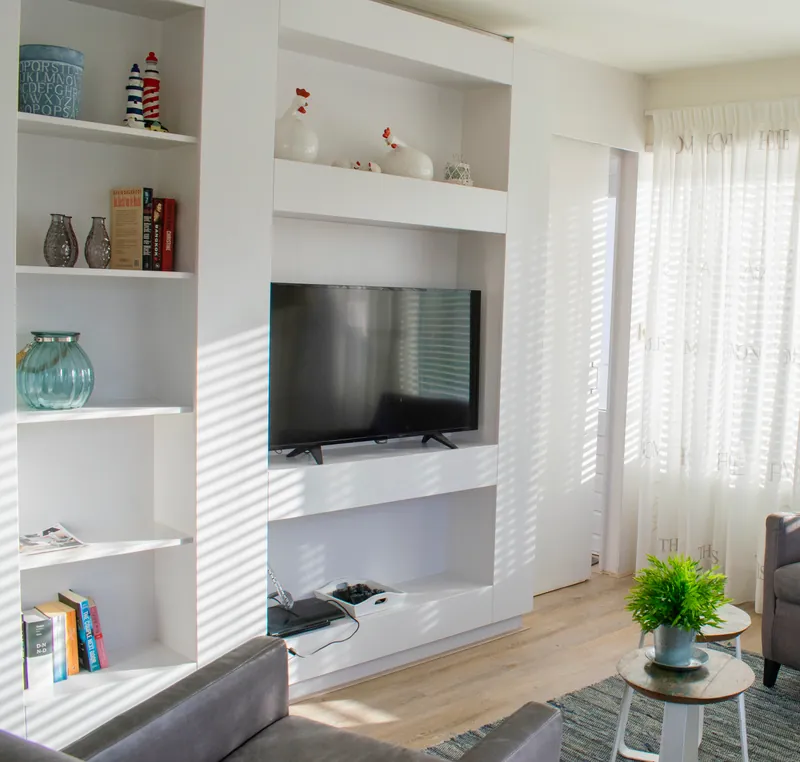 De woonkamer vanaf de andere kant: de tv in een witte wandkast met open schappen, en strepen zonlicht op de muur.